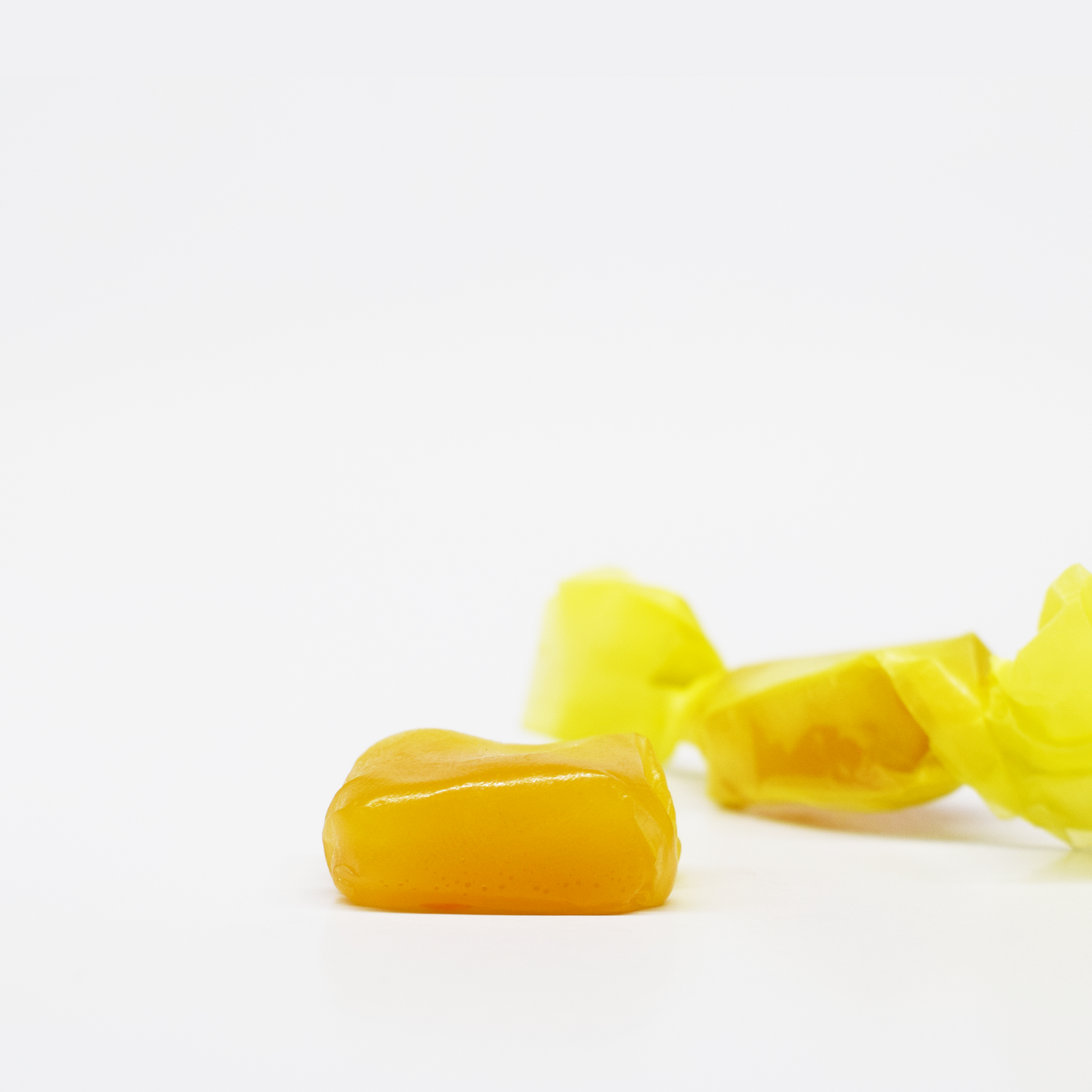 A single unwrapped Jolly Cannabis Pineapple Taffy (15mg THC, 15mg CBD) rests on a white surface, while another caramel in yellow wrapping appears blurred in the background, creating a clean and minimal look.