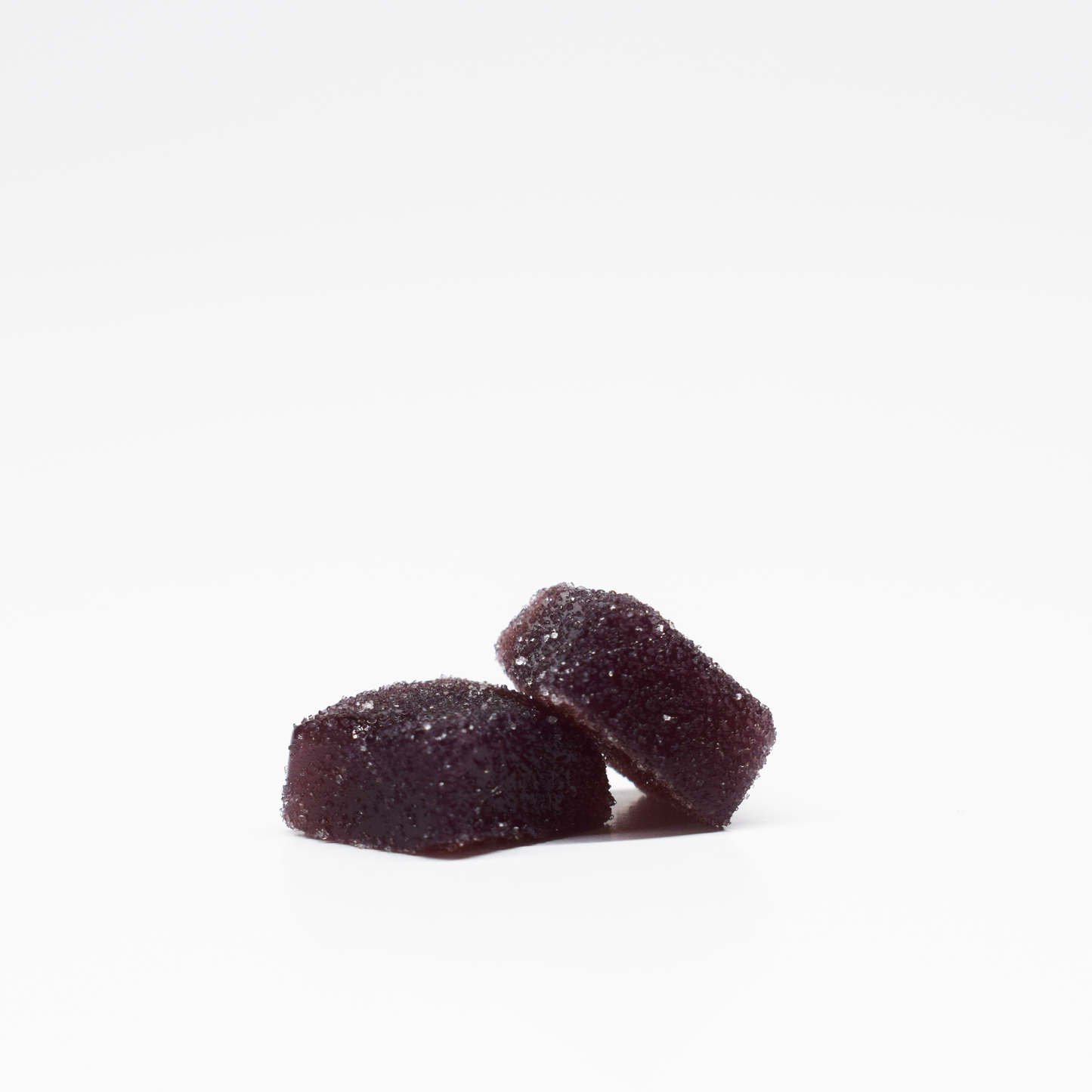 Two dark purple, sugar-coated Blueberry Raspberry Gummies 5mg THC, 125mg CBD from Jolly Cannabis are stacked on a white background. These candies have a soft, square shape with slightly rounded edges.