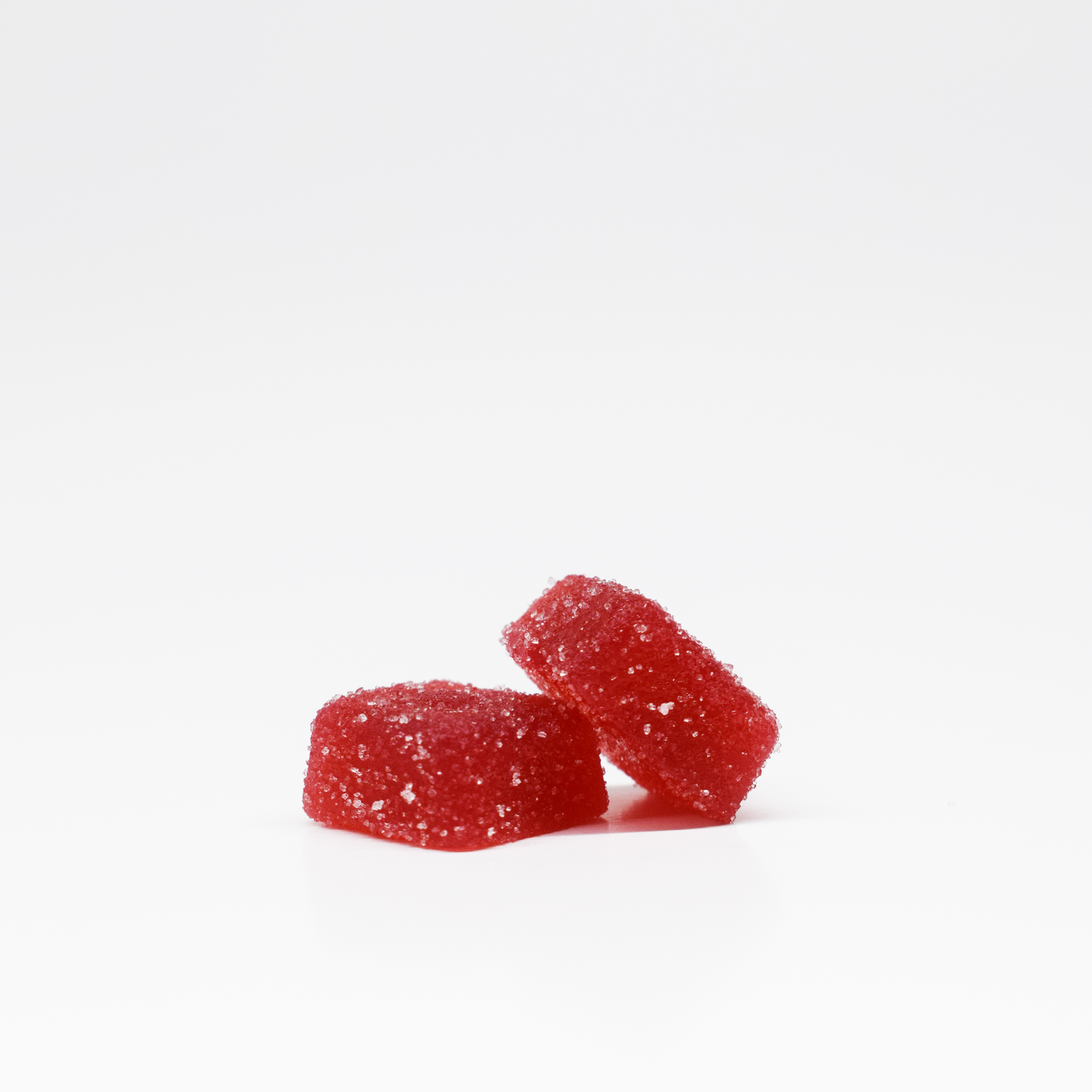 Two red Pomegranate Gummies (5mg THC, 125mg CBD) from Jolly Cannabis rest against each other on a white background. One lies flat while the other leans atop it, both coated in sparkling sugar crystals.
