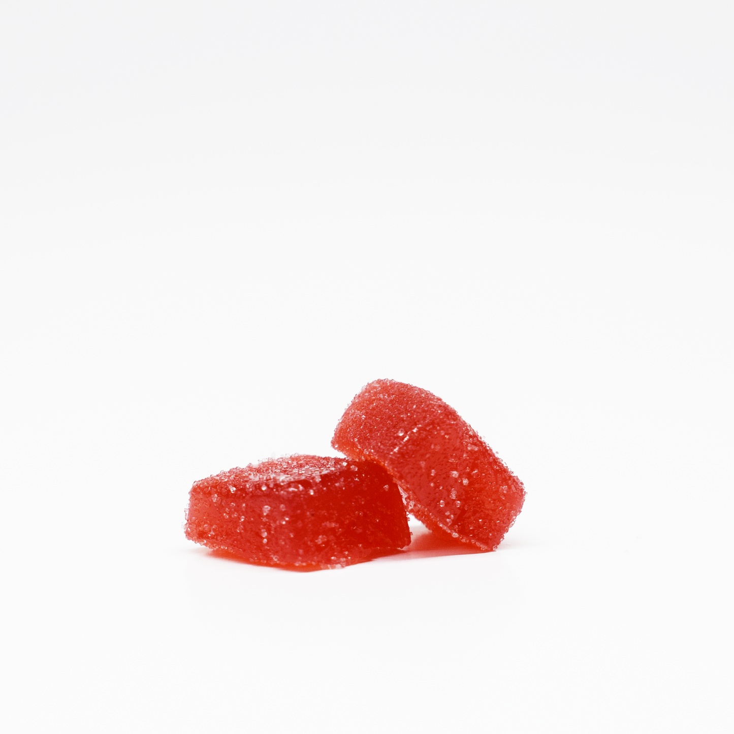 Two red, rectangular Strawberry Gummies 5mg THC, 125mg CBD by Jolly Cannabis are coated with sugar and placed on a white surface—one flat, one tilted—showcasing their vibrant color and sugary texture against the plain background.