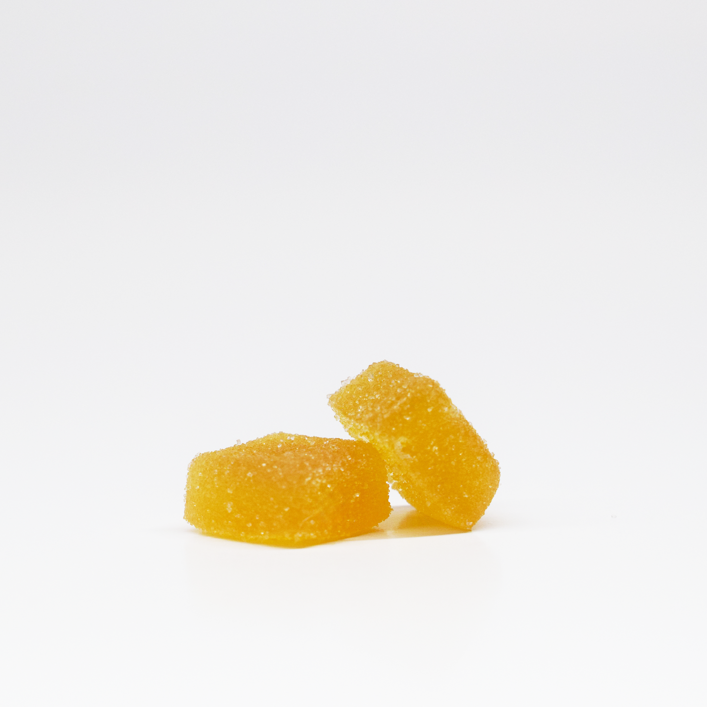 Two yellow White Peach Gummies 5mg THC, 125mg CBD by Jolly Cannabis sit on a white surface. One leans on the other, both with a soft, slightly translucent appearance and a rough sugar-coated texture.