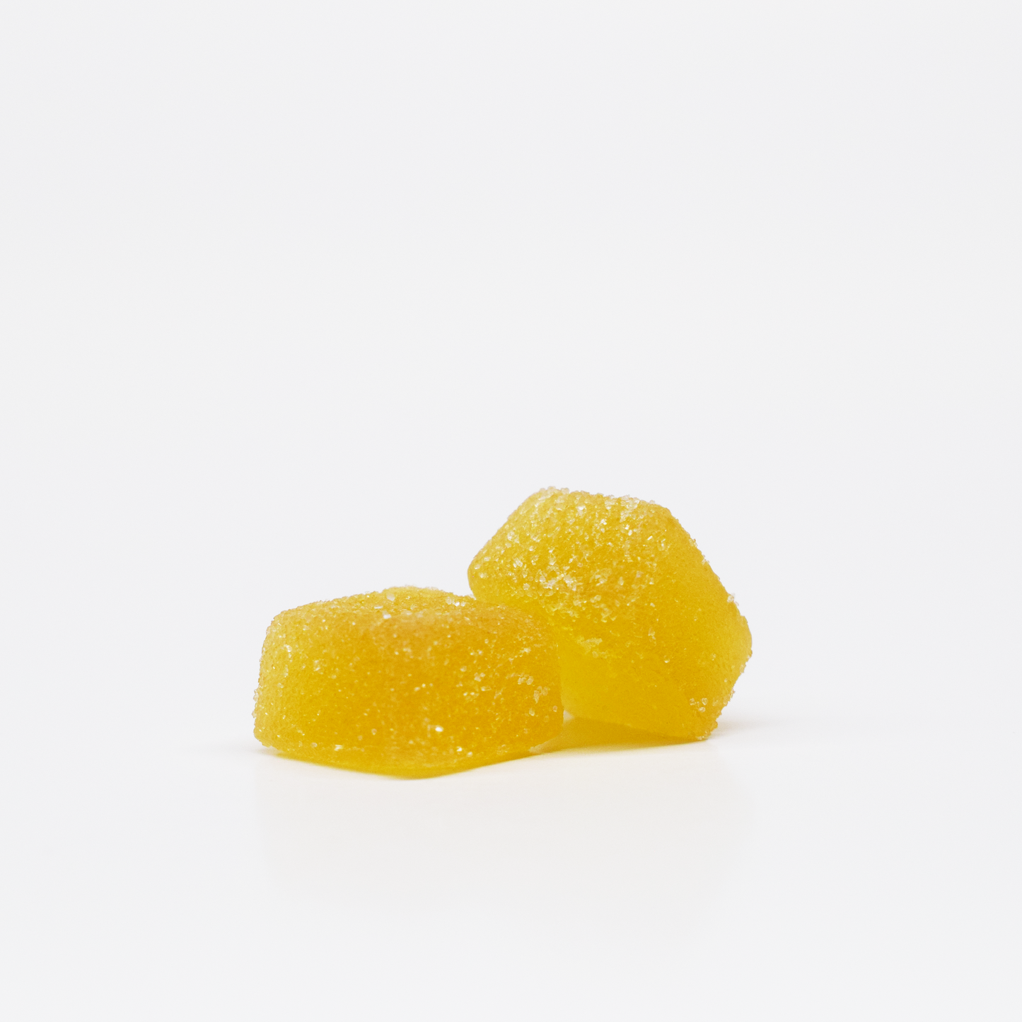 Two yellow Pineapple Gummies from Jolly Cannabis, each with 15mg THC and 15mg CBD, are stacked on a white background. Their soft, cube-like shapes have rounded edges and a sugar-coated, glistening surface.