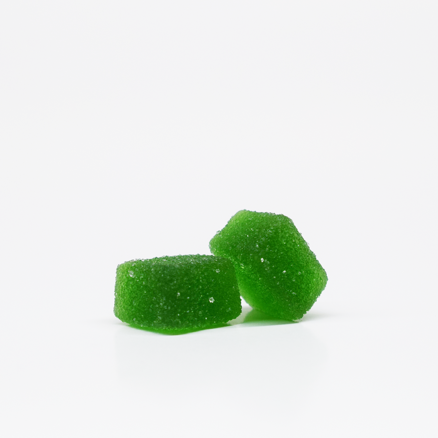 Two bright green Jolly Cannabis Sour Apple Gummies (15mg THC, 15mg CBD each) rest on a glossy white surface. Their smooth, slightly irregular shapes glisten with a light sugar crystal coating against a plain white background.