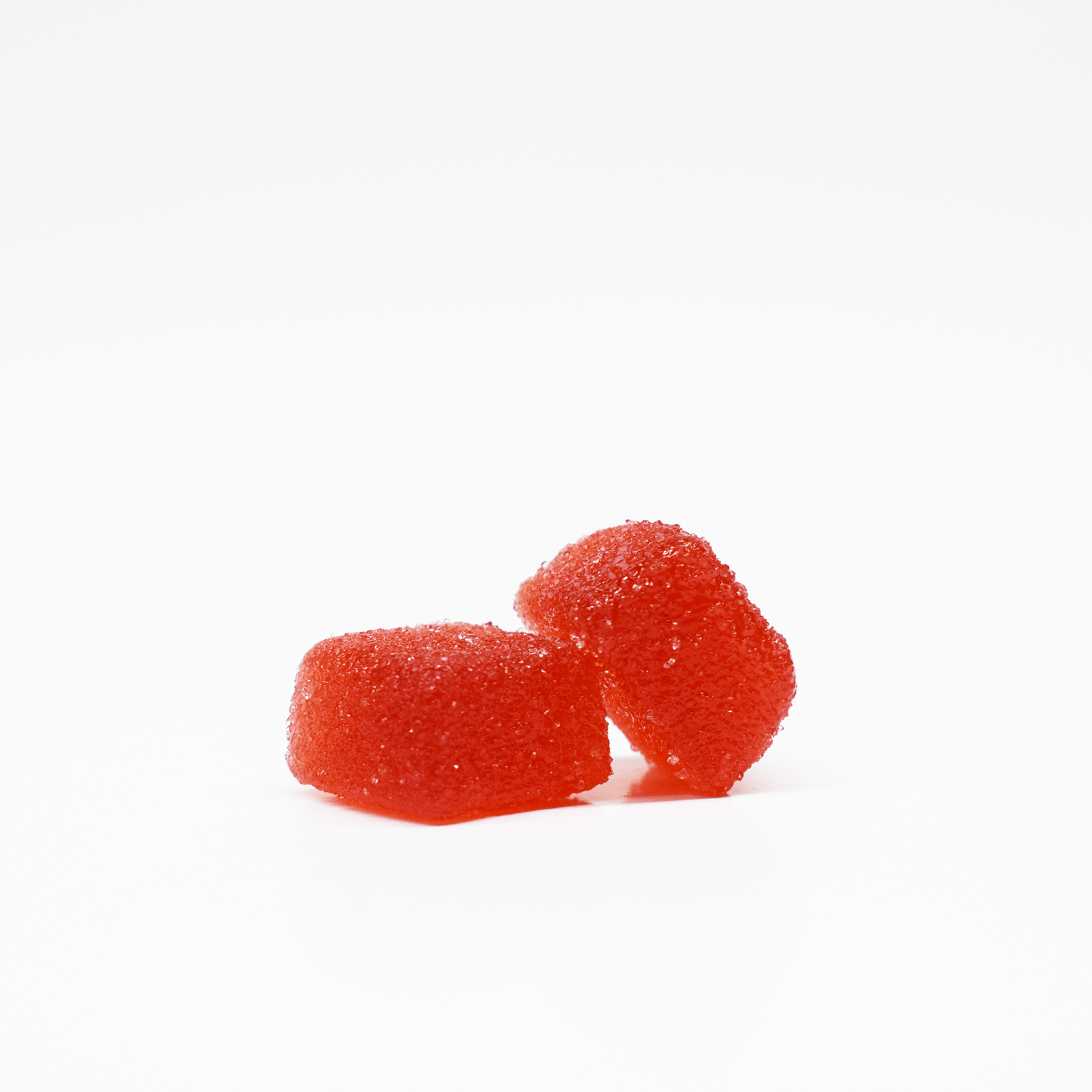 Two red, sugar-coated Jolly Cannabis Strawberry Watermelon Gummies (15mg THC, 15mg CBD each) sit on a plain white background—one upright and one leaning—showcasing their chewy texture and sparkling crystalline coating.