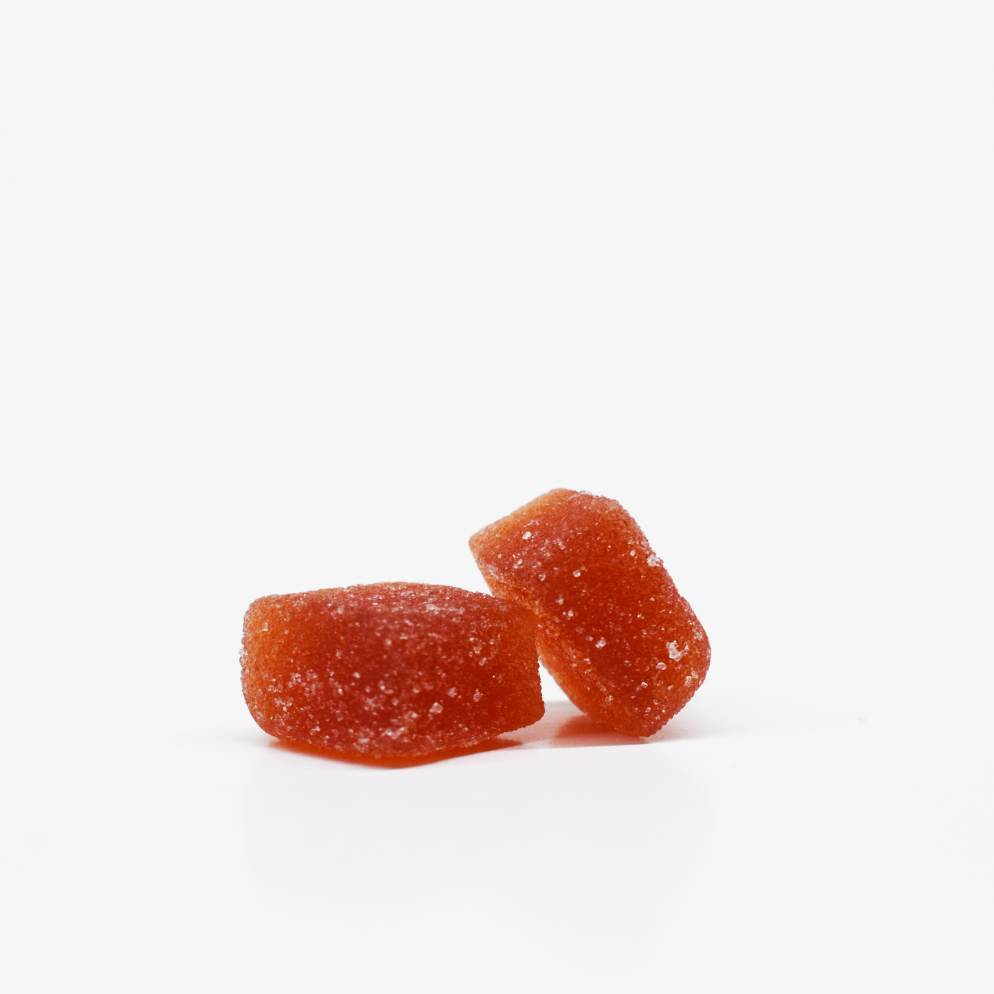 Two orange-red Tropical Gummies 15mg THC, 15mg CBD by Jolly Cannabis rest on a white background—one upright, one leaning. Their sugar-coated surfaces sparkle and reveal a slightly translucent fruit puree texture.
