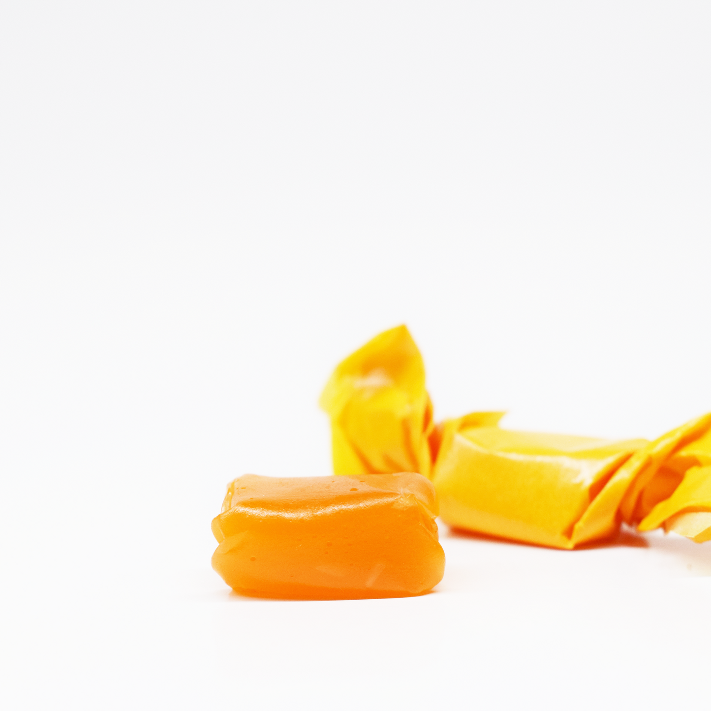 A close-up of two SimplyHemply Orange Passion Fruit Taffy chews (15mg THC, 15mg CBD) on a white background—one unwrapped to reveal its smooth surface, the other in yellow wax paper—positioned at the bottom right of the image.