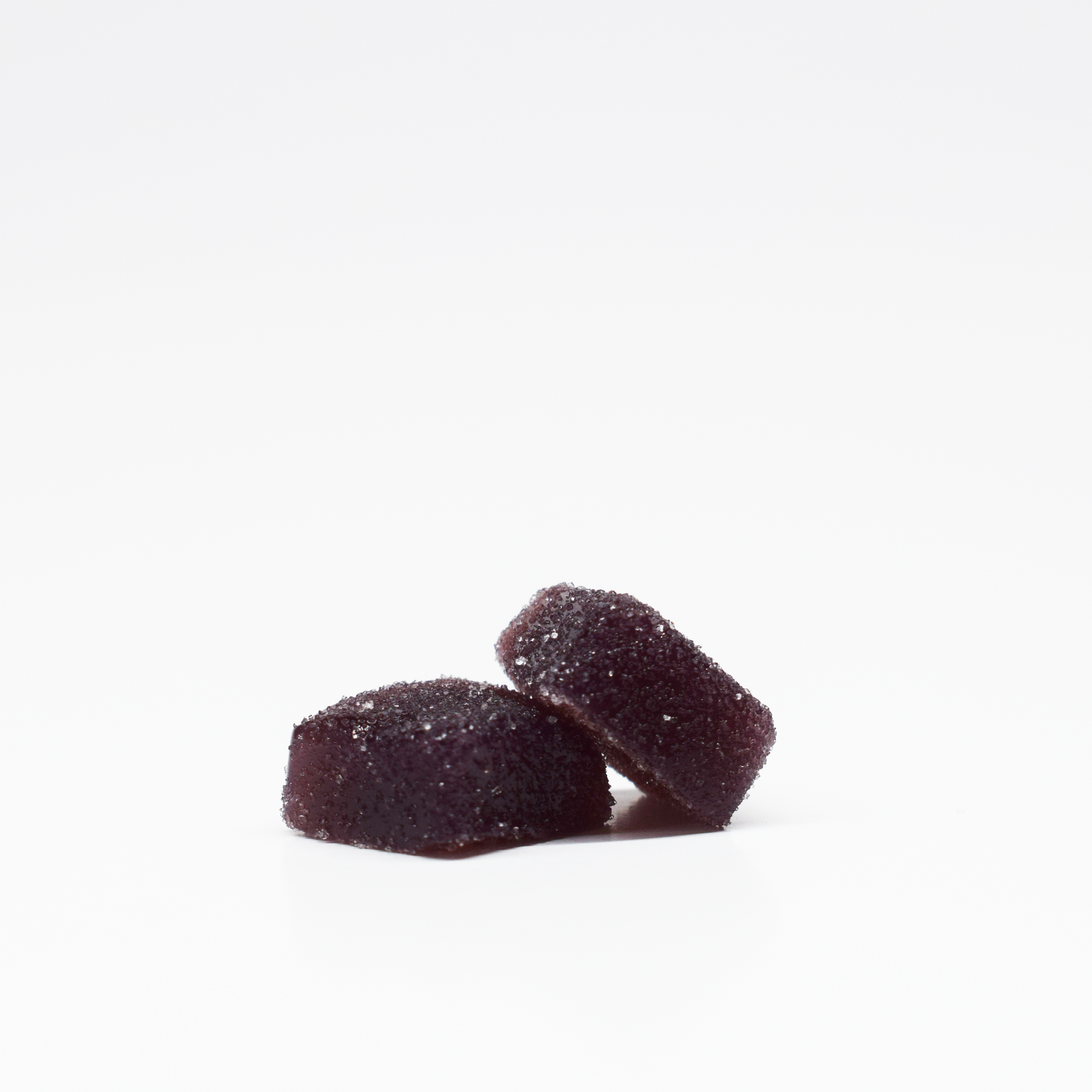Two dark purple, sugar-coated Blueberry Raspberry Gummies 5mg THC, 125mg CBD from Jolly Cannabis are stacked on a white background. These candies have a soft, square shape with slightly rounded edges.