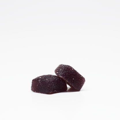 Two dark purple, sugar-coated Blueberry Raspberry Gummies 5mg THC, 125mg CBD from Jolly Cannabis are stacked on a white background. These candies have a soft, square shape with slightly rounded edges.