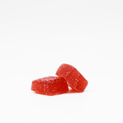 Two red, rectangular Strawberry Gummies 5mg THC, 125mg CBD by Jolly Cannabis are coated with sugar and placed on a white surface—one flat, one tilted—showcasing their vibrant color and sugary texture against the plain background.
