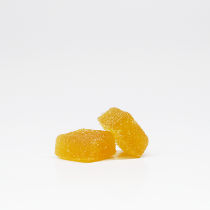 Two yellow White Peach Gummies 5mg THC, 125mg CBD by Jolly Cannabis sit on a white surface. One leans on the other, both with a soft, slightly translucent appearance and a rough sugar-coated texture.