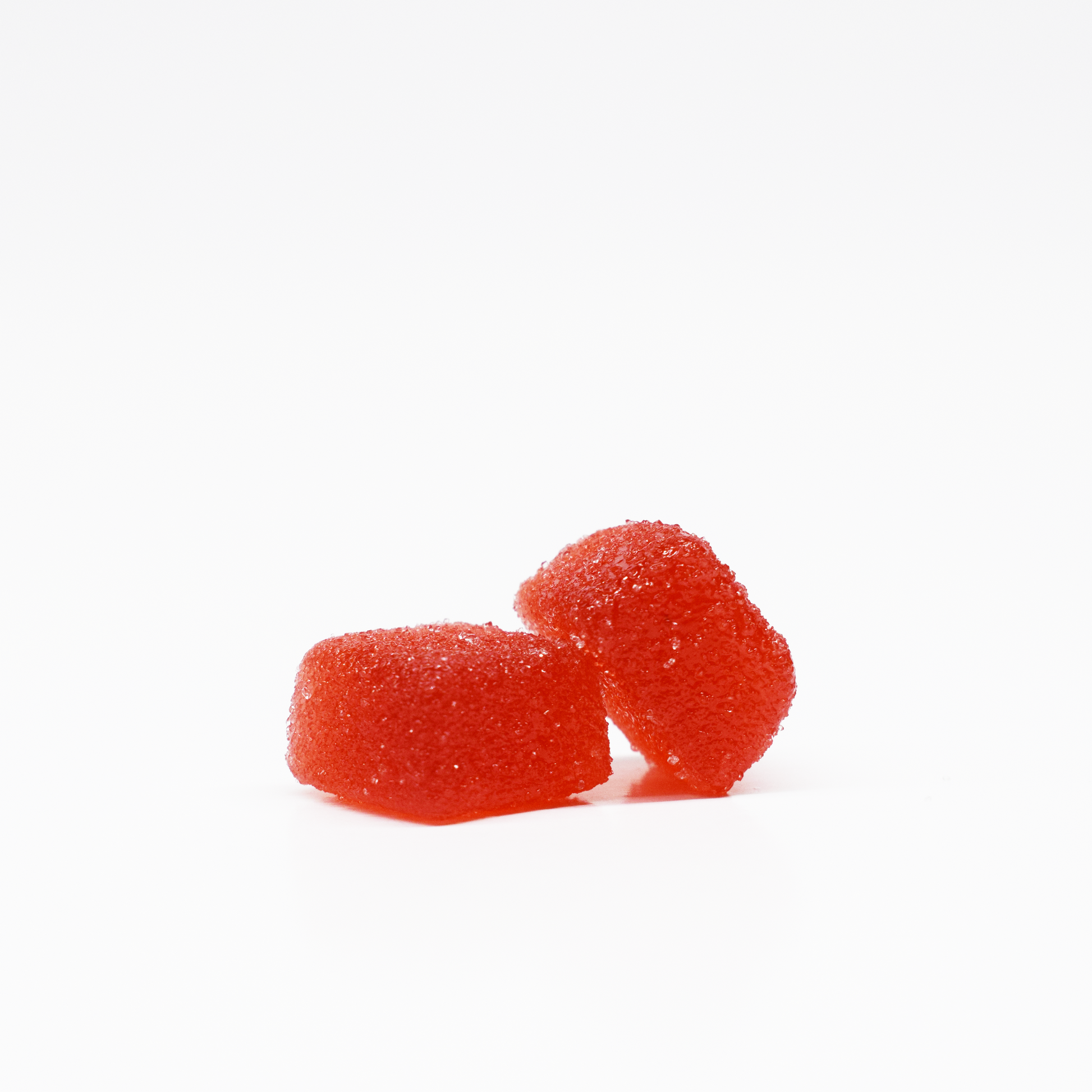 Two red, sugar-coated Jolly Cannabis Strawberry Watermelon Gummies (15mg THC, 15mg CBD each) sit on a plain white background—one upright and one leaning—showcasing their chewy texture and sparkling crystalline coating.